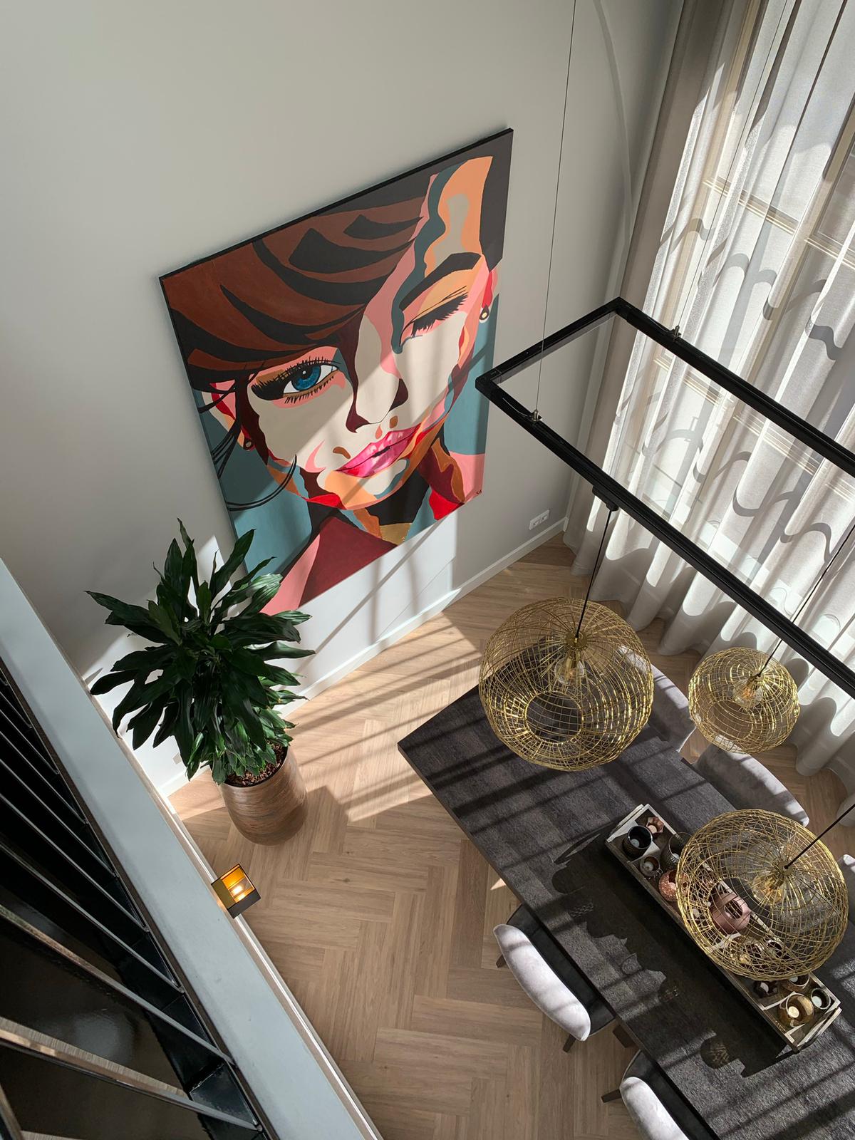 Groot kleurrijk vrouwenportret aan de muur in een lichte woonkamer met visgraatvloer en gouden hanglampen