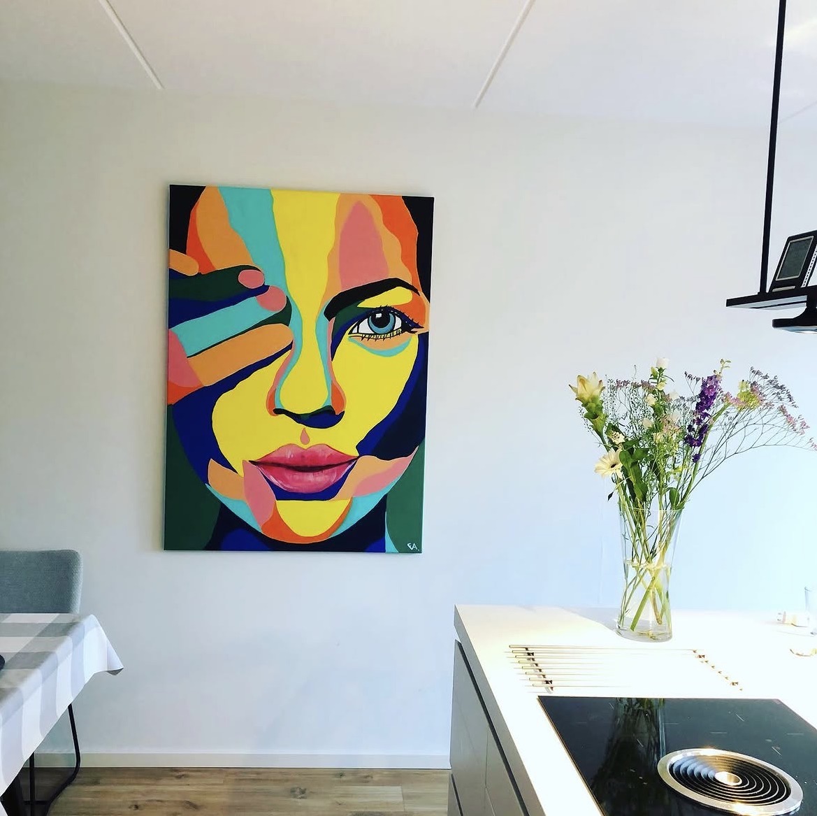 Colourful painted portrait of a woman holding her hand across her face, hanging on a white wall in a bright kitchen